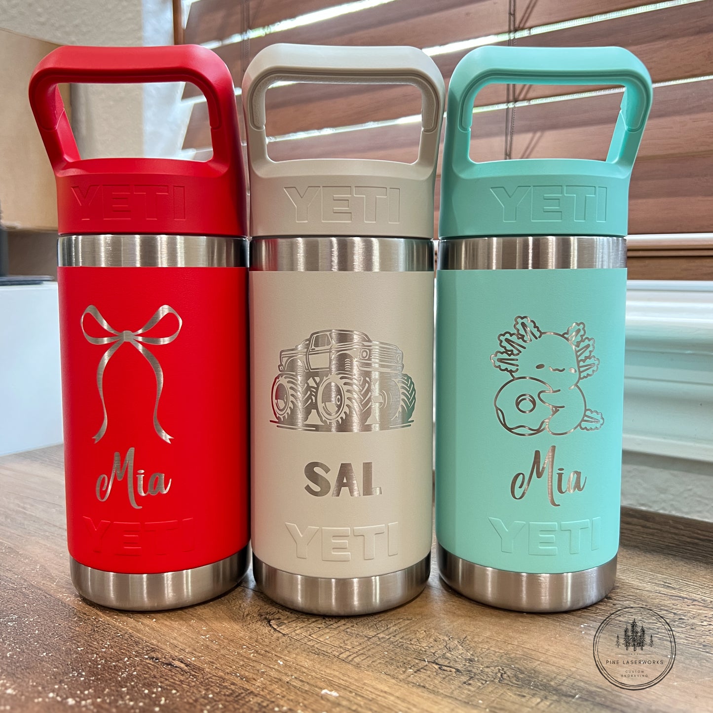 Three YETI insulated tumblers with carry handles are lined up on a wooden surface. The left tumbler is red with a bow engraving and the name 'Mia'.