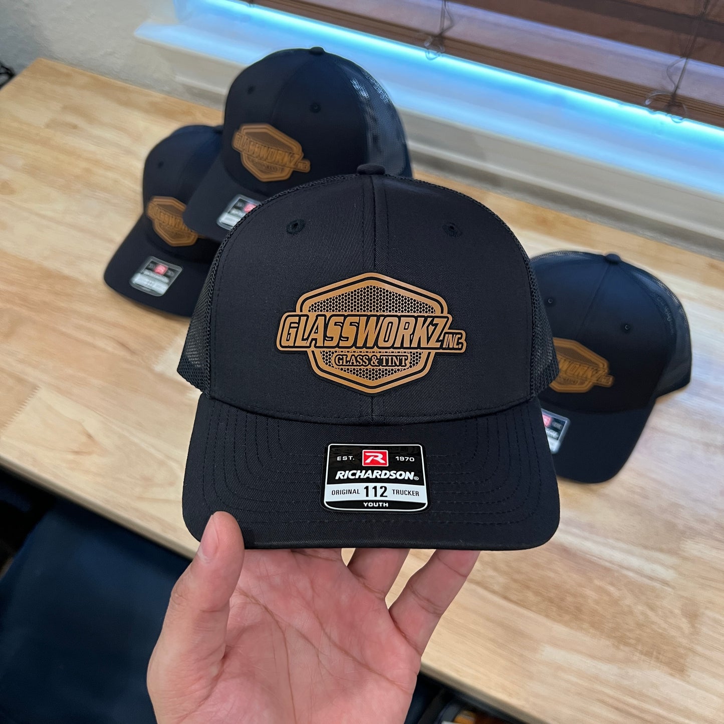 A hand holds a black Richardson 112 trucker hat with a gold and black 'GLASSWORKZ' patch on the front, with three more matching hats in the background on a wooden table.