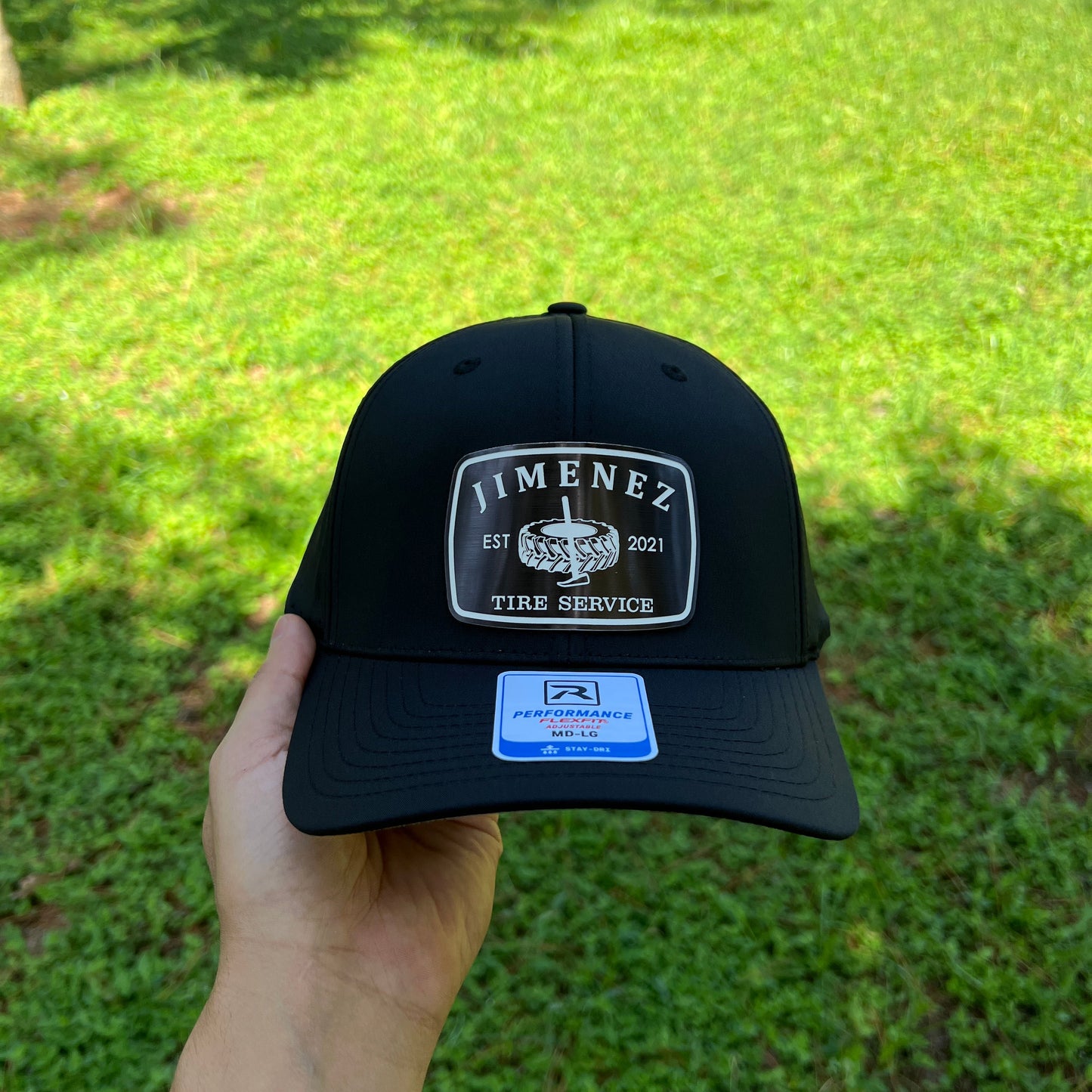 A black snapback cap held by a hand against a green grass background. The cap features a silver and black 'JIMENEZ' patch on the front and a blue and white sticker on the side.
