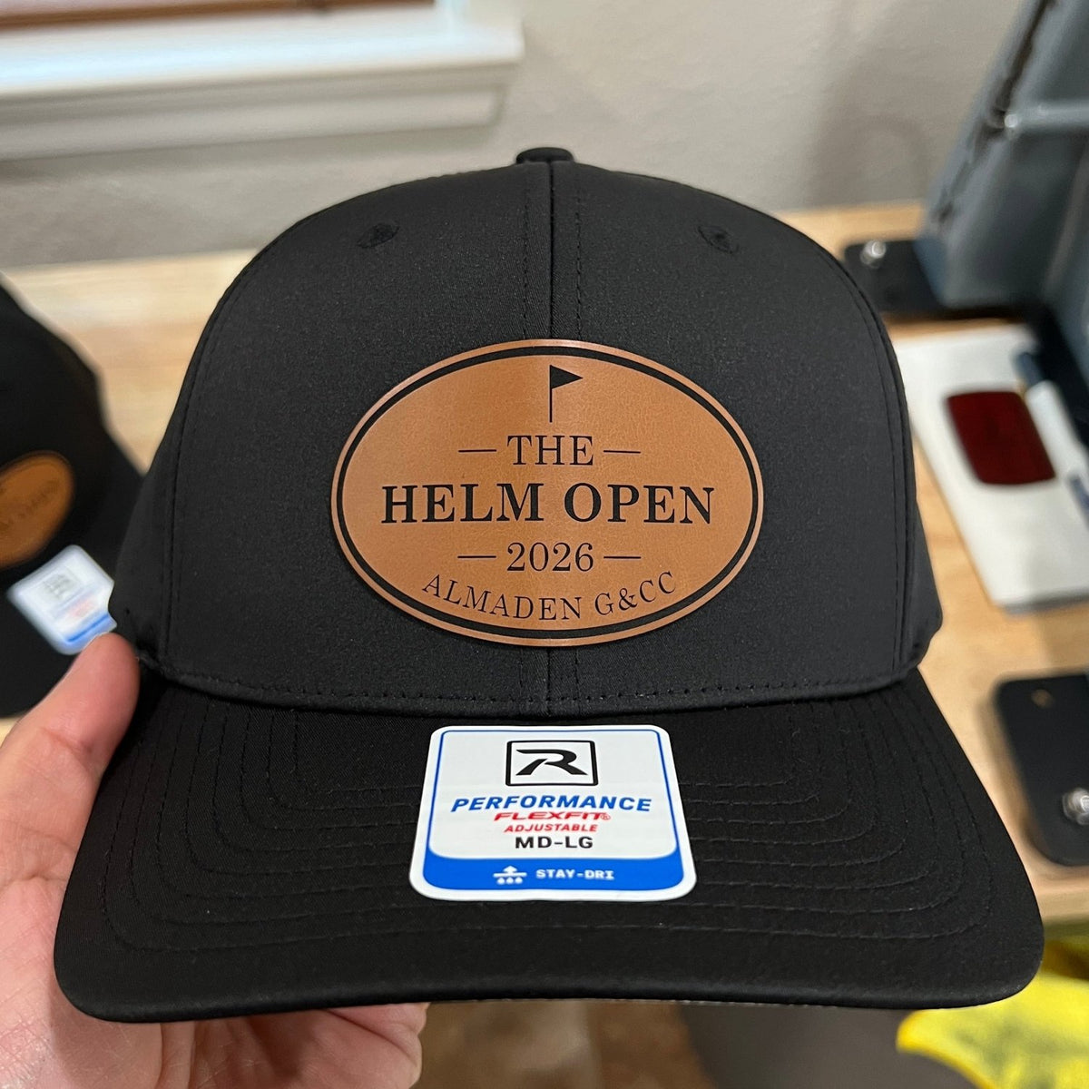 A black baseball cap held by a hand, featuring a brown oval leather patch on the front that reads 'THE HELM OPEN 2026 ALMADEN G&CO'. A white and blue Performance brand sticker is attached to the brim.