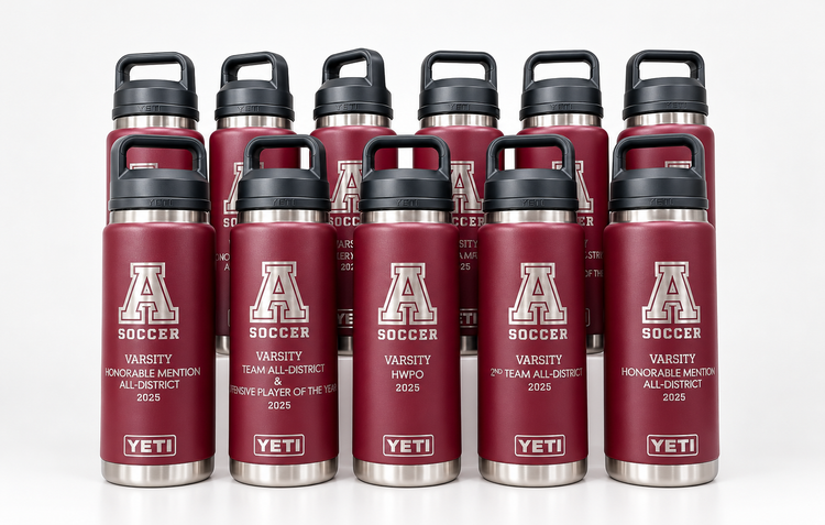 Six customized Yeti 26oz vacuum-insulated stainless steel water bottles with black lids, arranged in two rows on a wooden surface against a light gray wall. The bottles are maroon with silver graphics featuring the letter 'A' and 'SOCCER'.