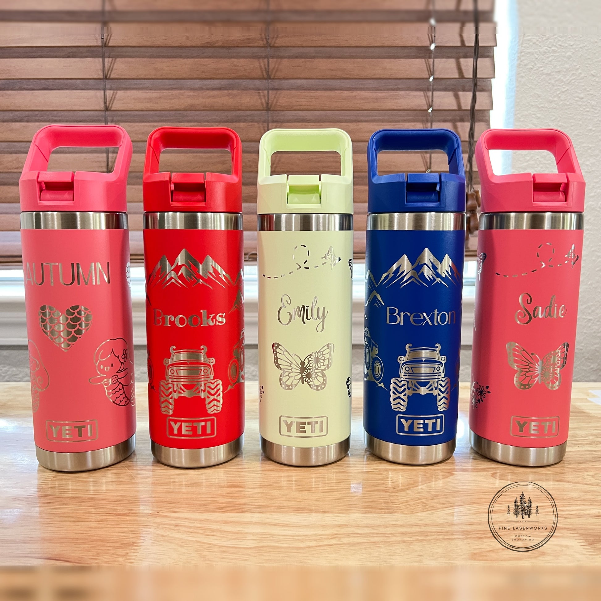 Five YETI travel tumblers with flip-top lids are lined up on a wooden table in front of window blinds. The tumblers come in pink, red, light green, blue, and coral colors, each decorated with a different travel-themed graphic and text including 'Autumn', 'Backroads', 'Emily', 'Brexton', and 'Sadie'.