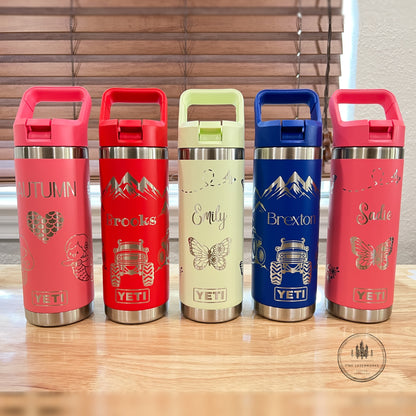Five YETI travel tumblers with flip-top lids are lined up on a wooden table in front of window blinds. The tumblers come in pink, red, light green, blue, and coral colors, each decorated with a different travel-themed graphic and text including 'Autumn', 'Backroads', 'Emily', 'Brexton', and 'Sadie'.