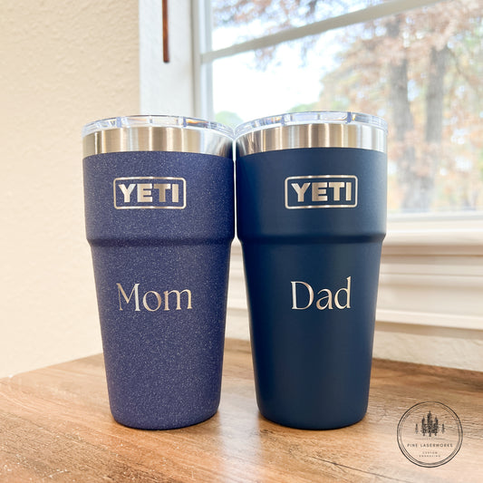 Two YETI tumblers labeled 'Mom' and 'Dad' on a wooden surface with a window in the background.