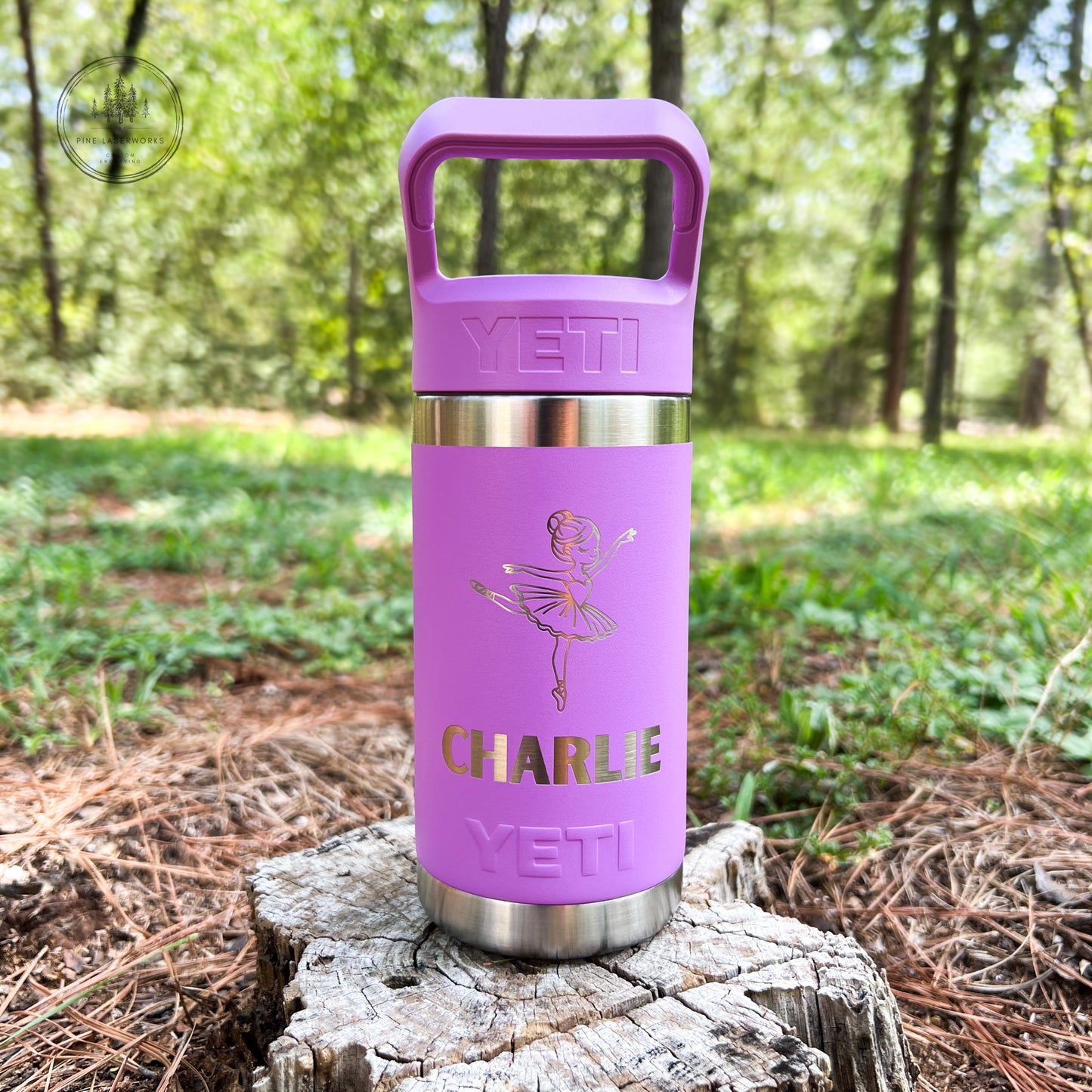 A purple YETI insulated tumbler with a handle lid, featuring a gold foil ballerina design and the name 'CHARLIE', displayed on a tree stump in a forest setting.