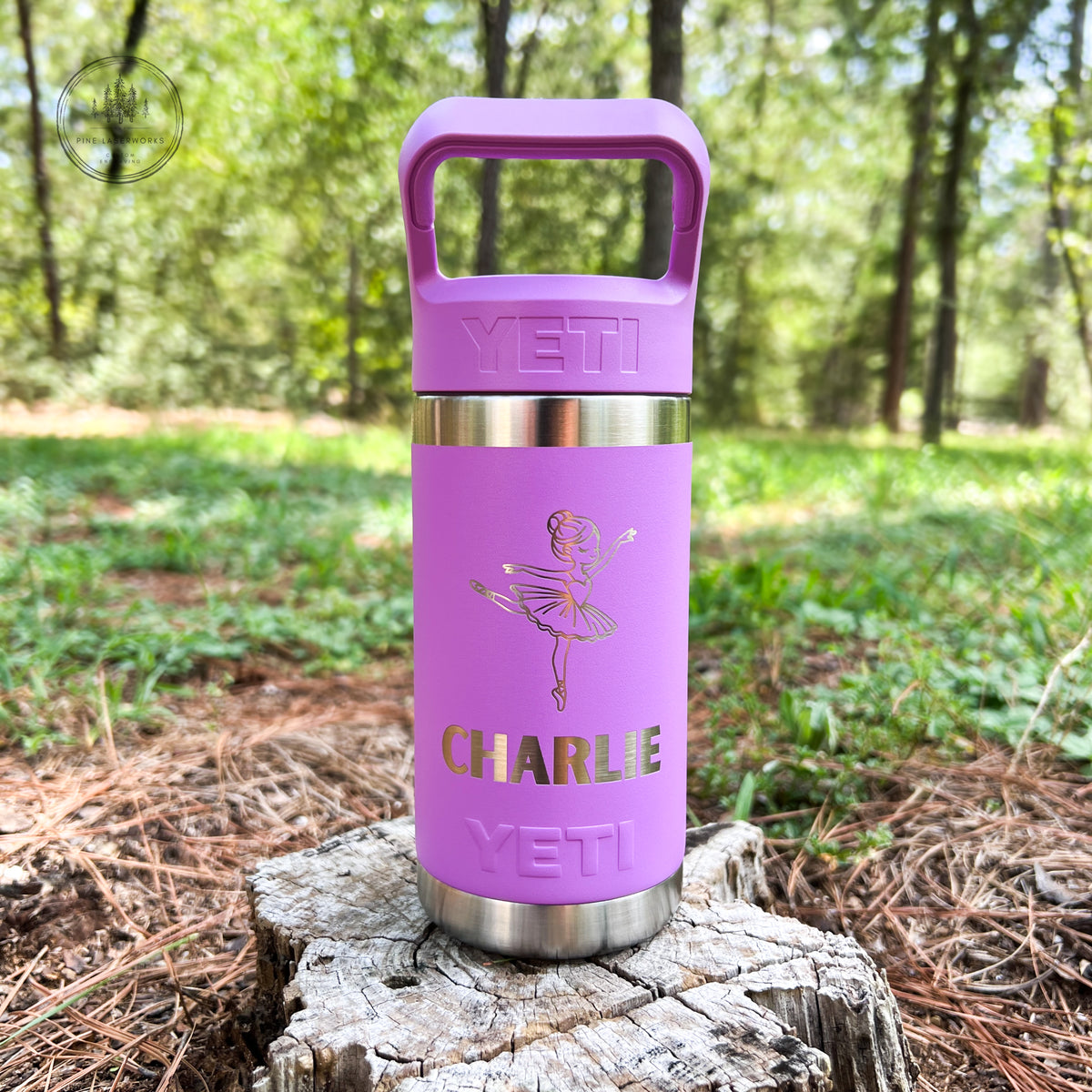 A purple YETI insulated tumbler with a handle lid, featuring a gold foil ballerina design and the name 'CHARLIE', displayed on a tree stump in a forest setting.