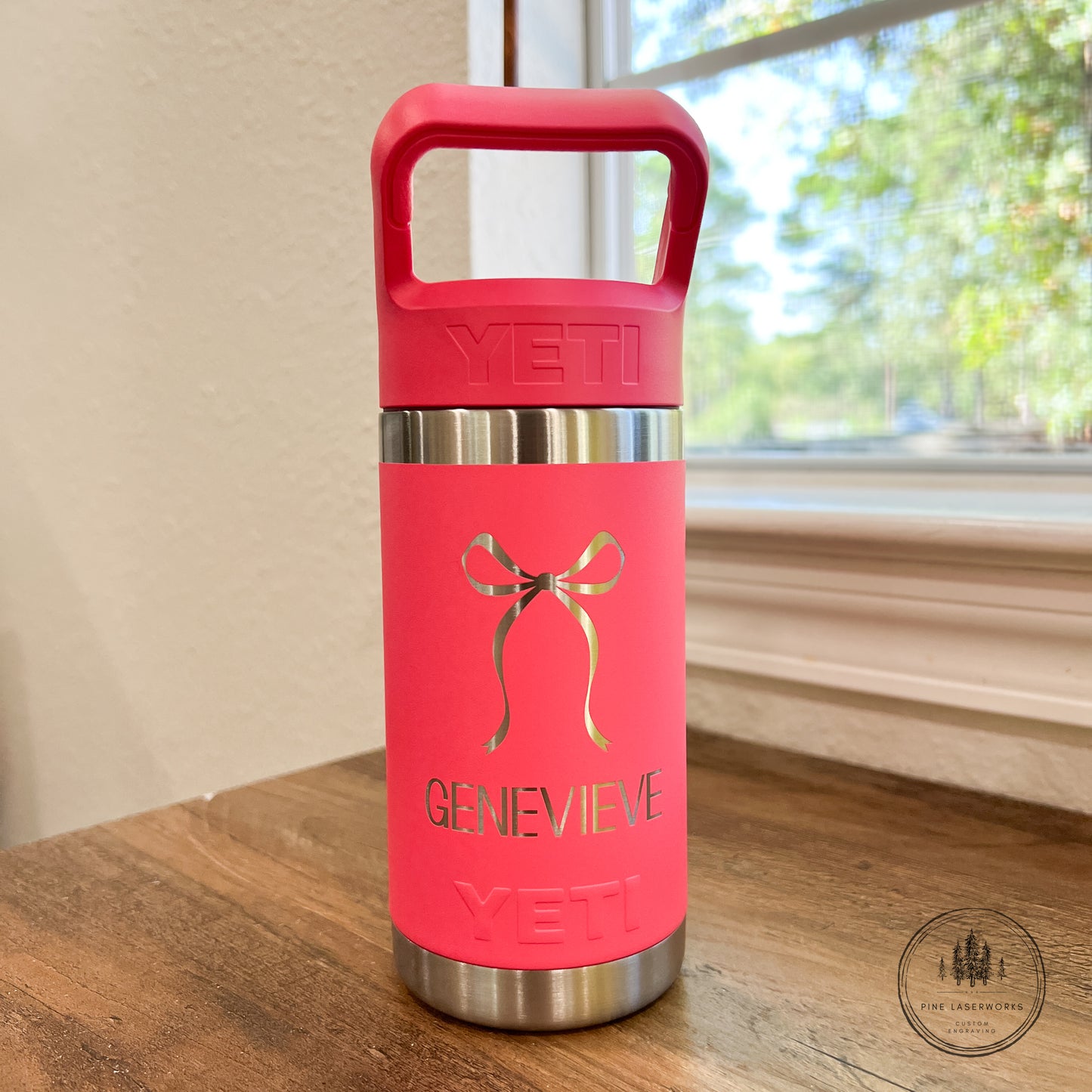 Pink YETI water bottle with 'Genevieve' on a wooden surface near a window.