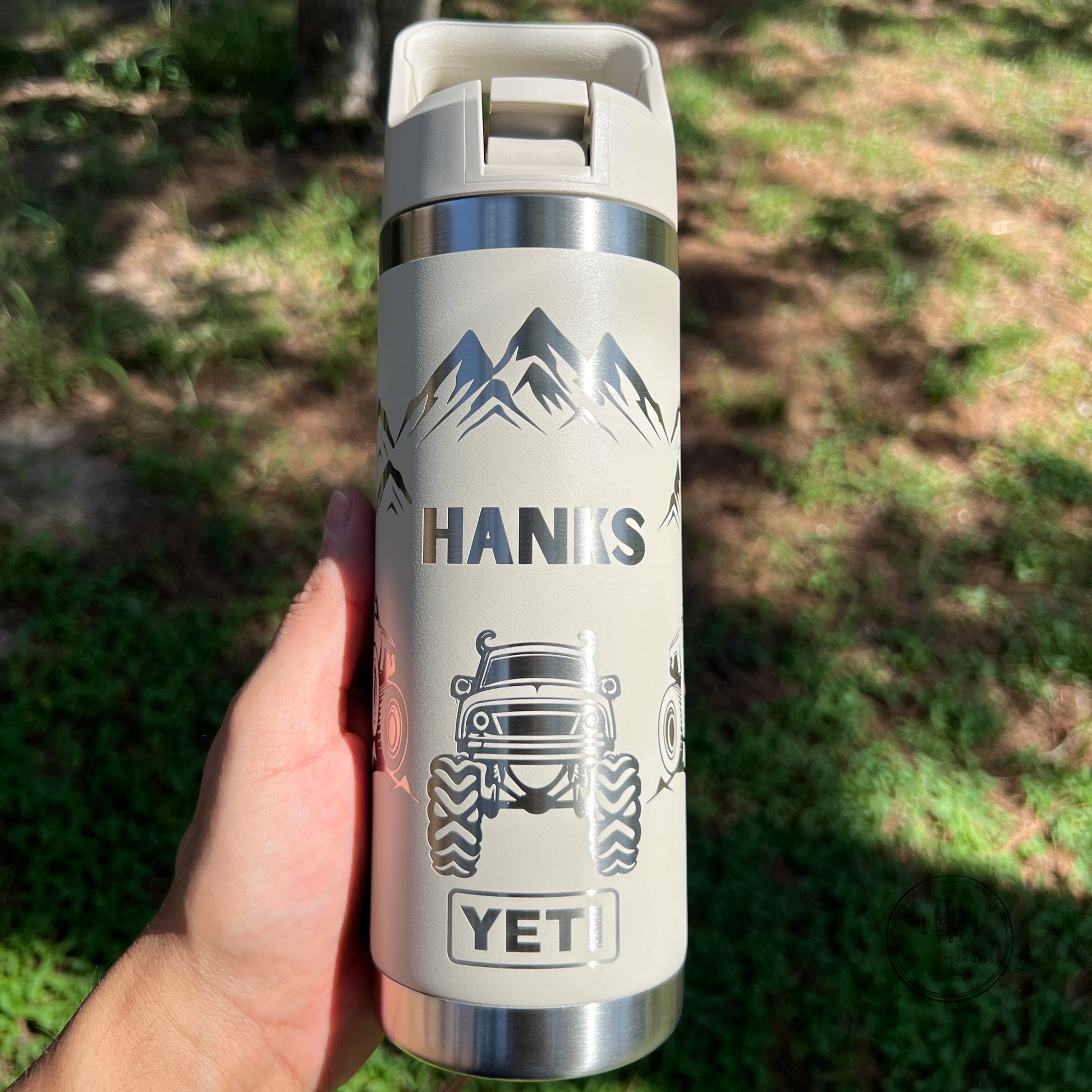 A hand holding a cream-colored Cape Taupe Yeti insulated water bottle outdoors. The bottle features a black graphic of a mountain range, the word 'HANKS' in a bold sans-serif font, a black monster truck illustration, and a 'YETI' logo at the bottom.