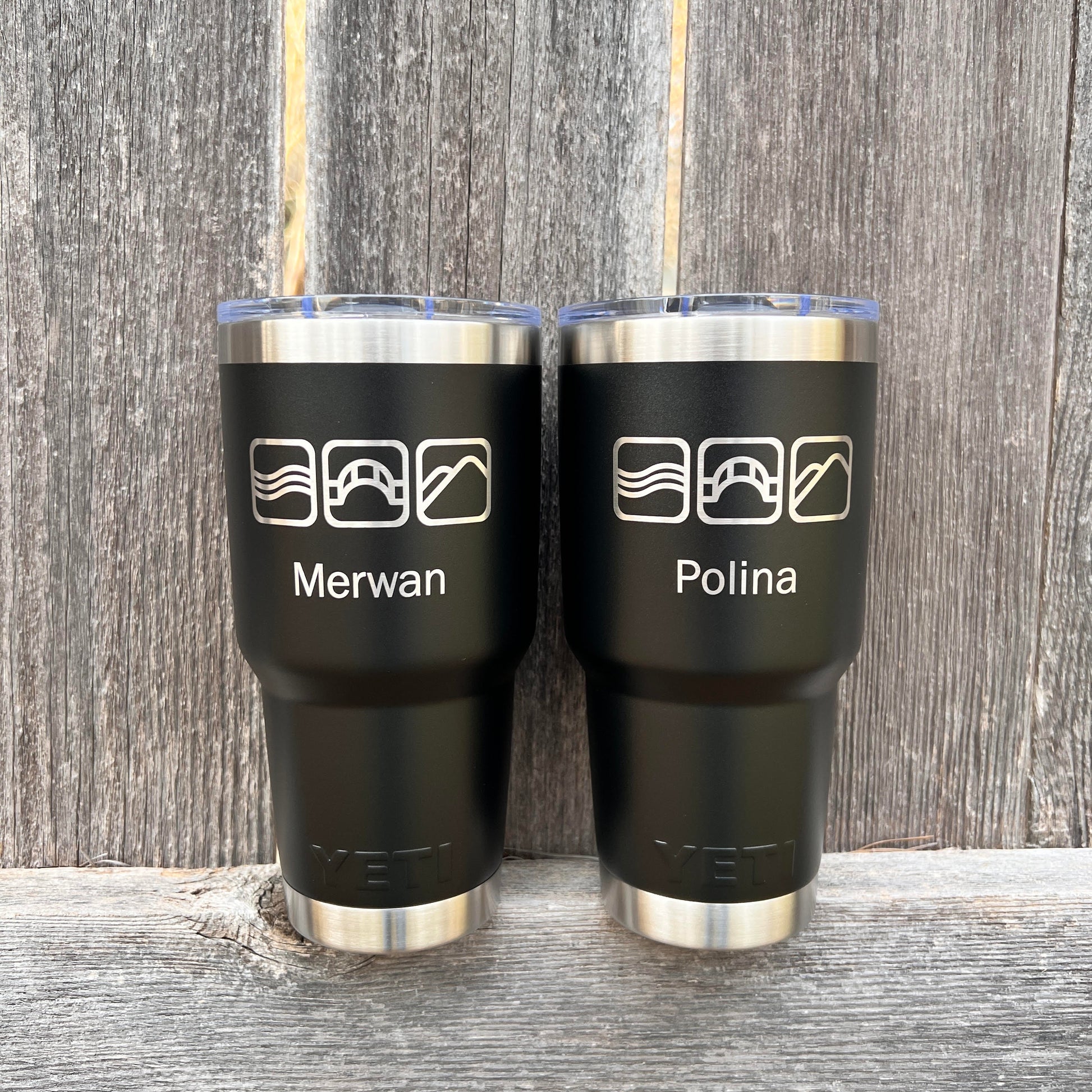Two black tumblers with names engraved on them, placed against a wooden background.