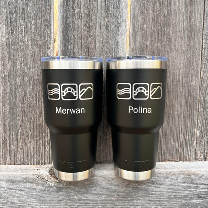 Two black tumblers with names engraved on them, placed against a wooden background.