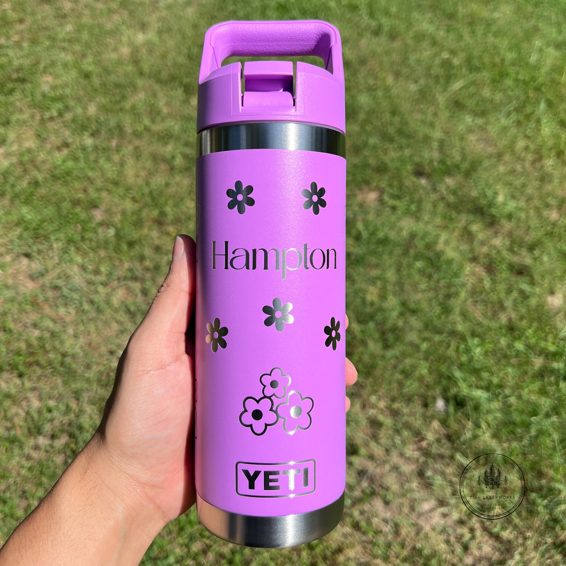 A hand holding a purple YETI insulated travel tumbler with a matching purple lid, featuring the name 'Hampton' and flower graphics, set against a blurred green grass background.