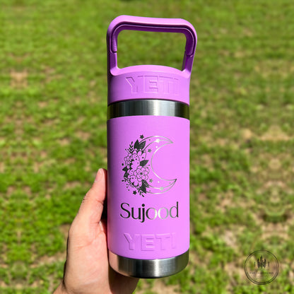 A hand holding a purple YETI insulated water bottle outdoors against a blurred green grass background. The bottle has a matching purple handle, stainless steel accents, and a graphic of a crescent moon and flowers with the text 'Sujood'.