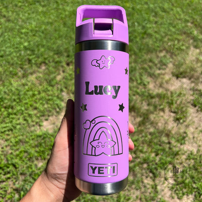 A hand holds a purple insulated water bottle outdoors against a blurred green grass background. The bottle has a matching purple lid, black text reading 'Lucy', and a black outlined rainbow and star design, with YETI branding visible near the bottom.