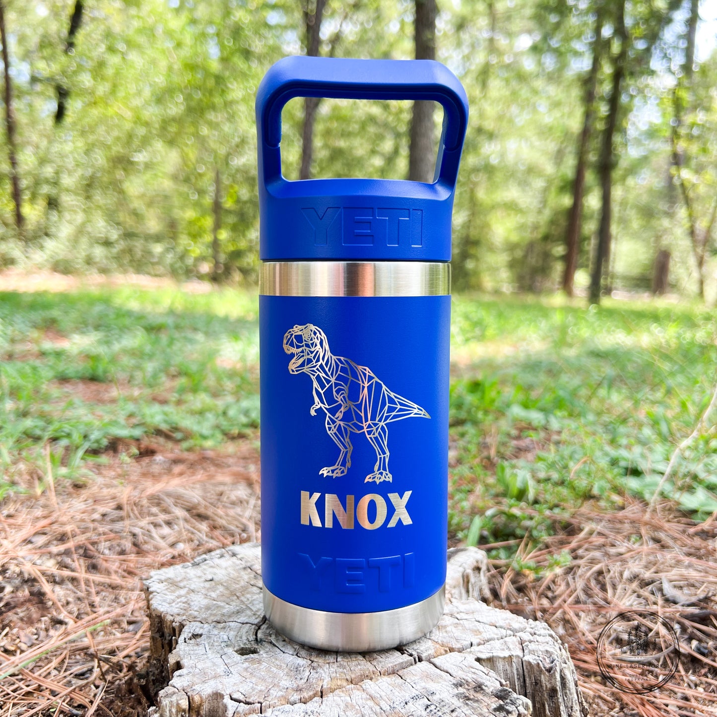 A blue YETI insulated tumbler with a handle, featuring a laser-engraved white dinosaur skeleton design and the name 'KNOX' in gold, standing on a tree stump in a forest setting.