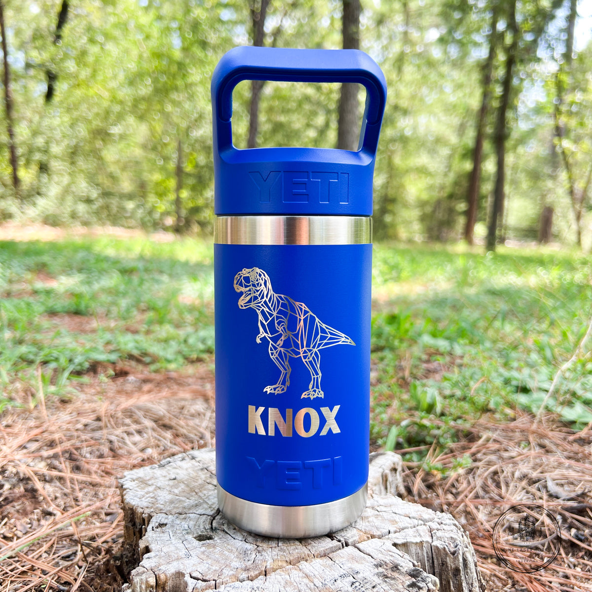 A blue YETI insulated tumbler with a handle, featuring a laser-engraved white dinosaur skeleton design and the name 'KNOX' in gold, standing on a tree stump in a forest setting.