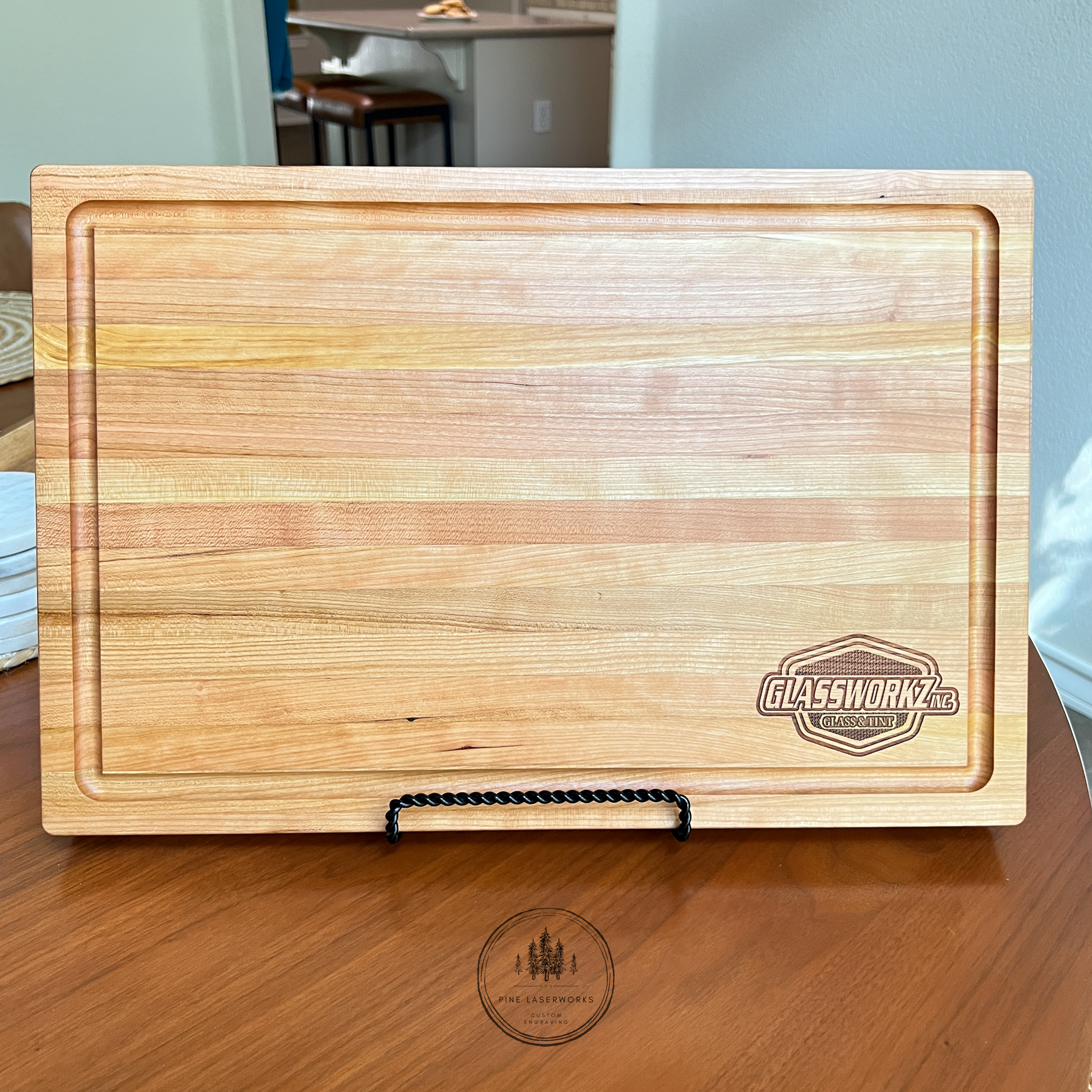 A rectangular wooden serving board with a recessed juice groove cut out on the perimeter, on a polished wooden table. The background shows a softly blurred interior room with a wall and a circular 'Pine Laserworks custom engraving' logo watermark near the bottom.