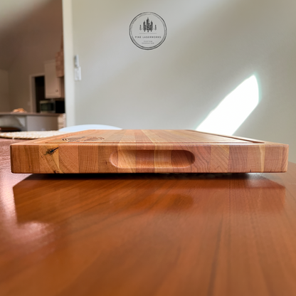 A rectangular wooden serving board with a recessed handle cutout on one side, photographed from a low angle on a polished wooden table. The background shows a softly blurred interior room with a wall and a circular 'Pine Laserworks custom engraving' logo watermark near the top.
