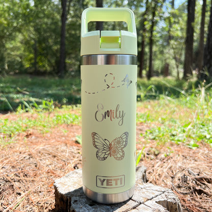 A grasshopper yellow-green YETI insulated tumbler with a matching lid and carry handle, standing on a tree stump outdoors. The tumbler is personalized with the name 'Emily' in cursive, a butterfly graphic, and a YETI logo near the bottom.