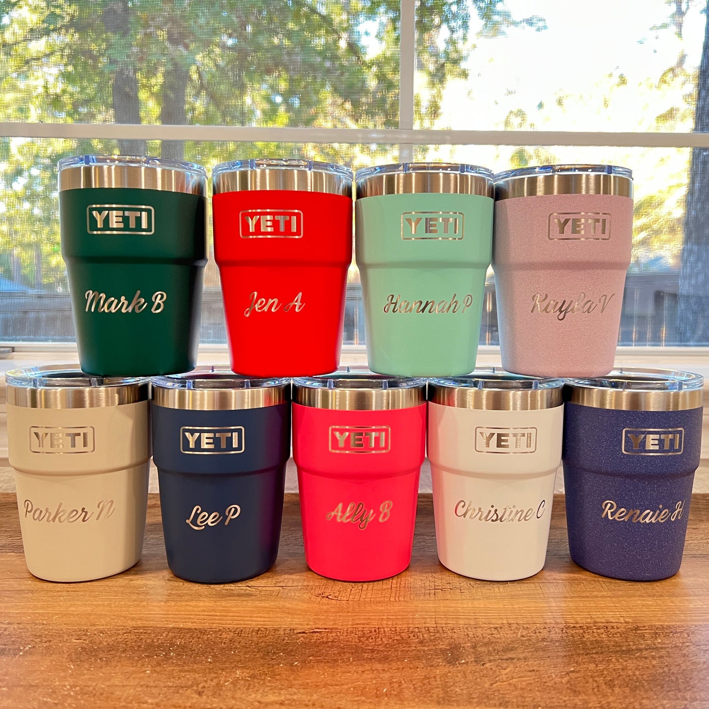 9 YETI insulated tumblers with stainless steel lids are arranged in two rows on a wooden table in front of a window. Each tumbler has a different color and a custom name engraved on the front.