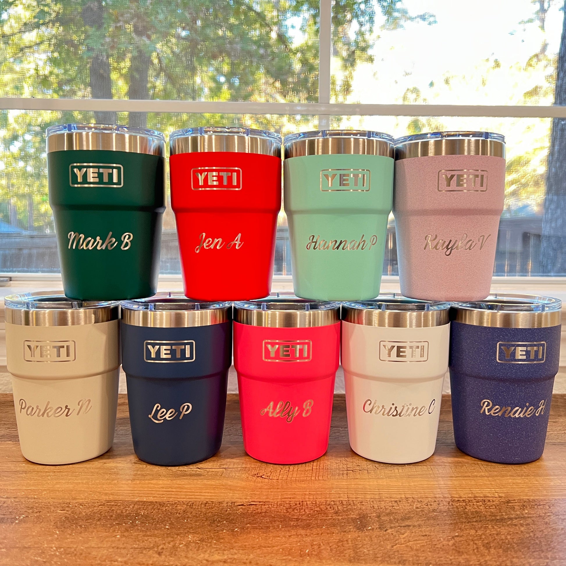 9 YETI insulated tumblers with stainless steel lids are arranged in two rows on a wooden table in front of a window. Each tumbler has a different color and a custom name engraved on the front.