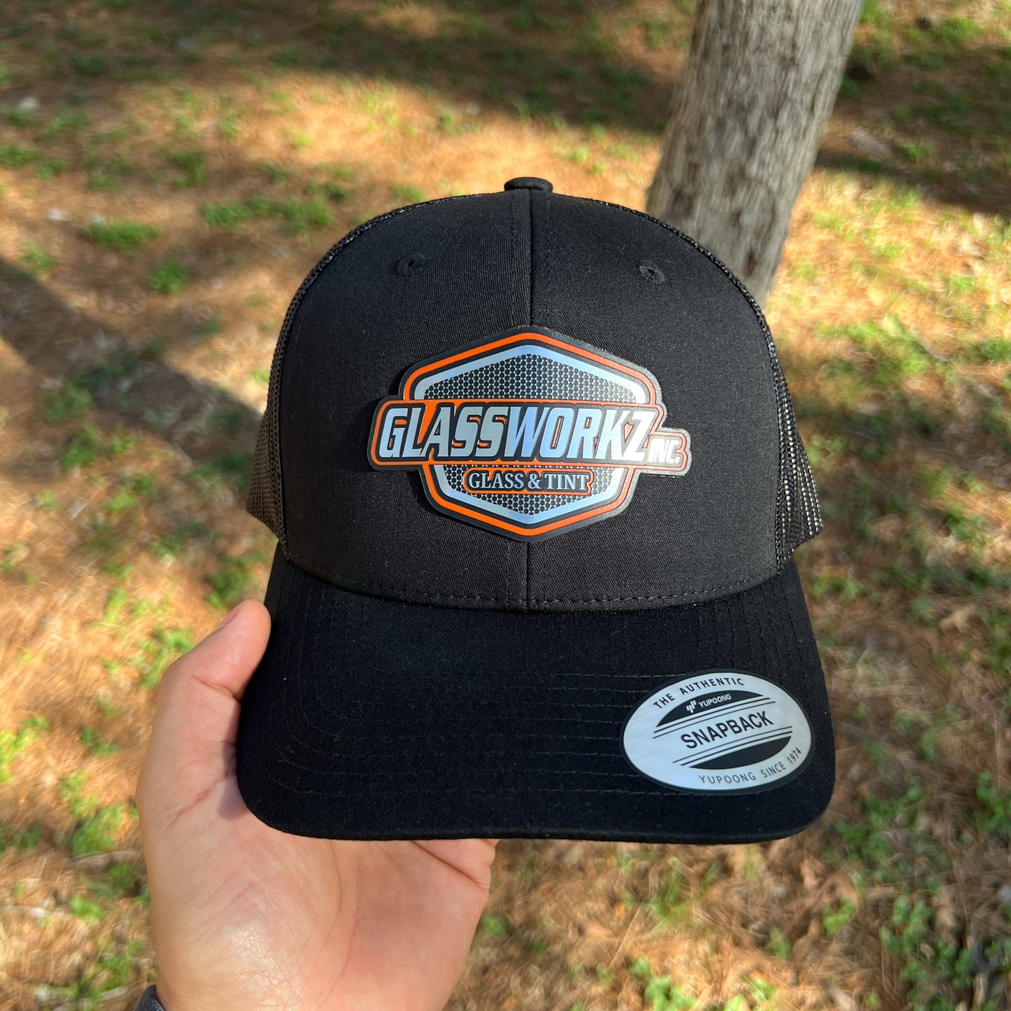 Black cap with Glassworks Glass & Tint logo held in front of a tree and grass background