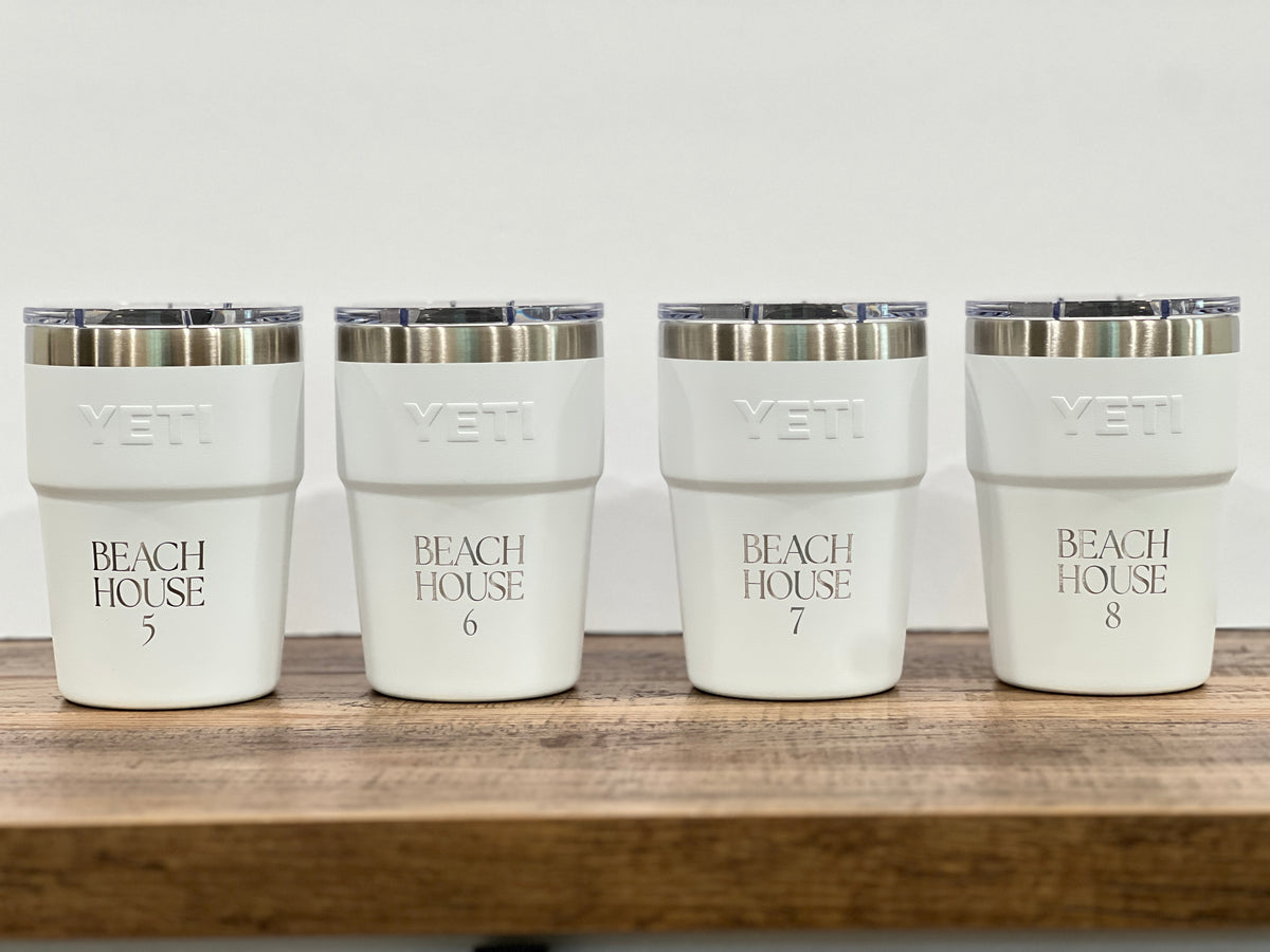 Four white YETI insulated tumblers with clear lids and silver rims, lined up on a wooden surface against a white background. Each cup is printed with 'BEACH HOUSE' and a number from 5 to 8.