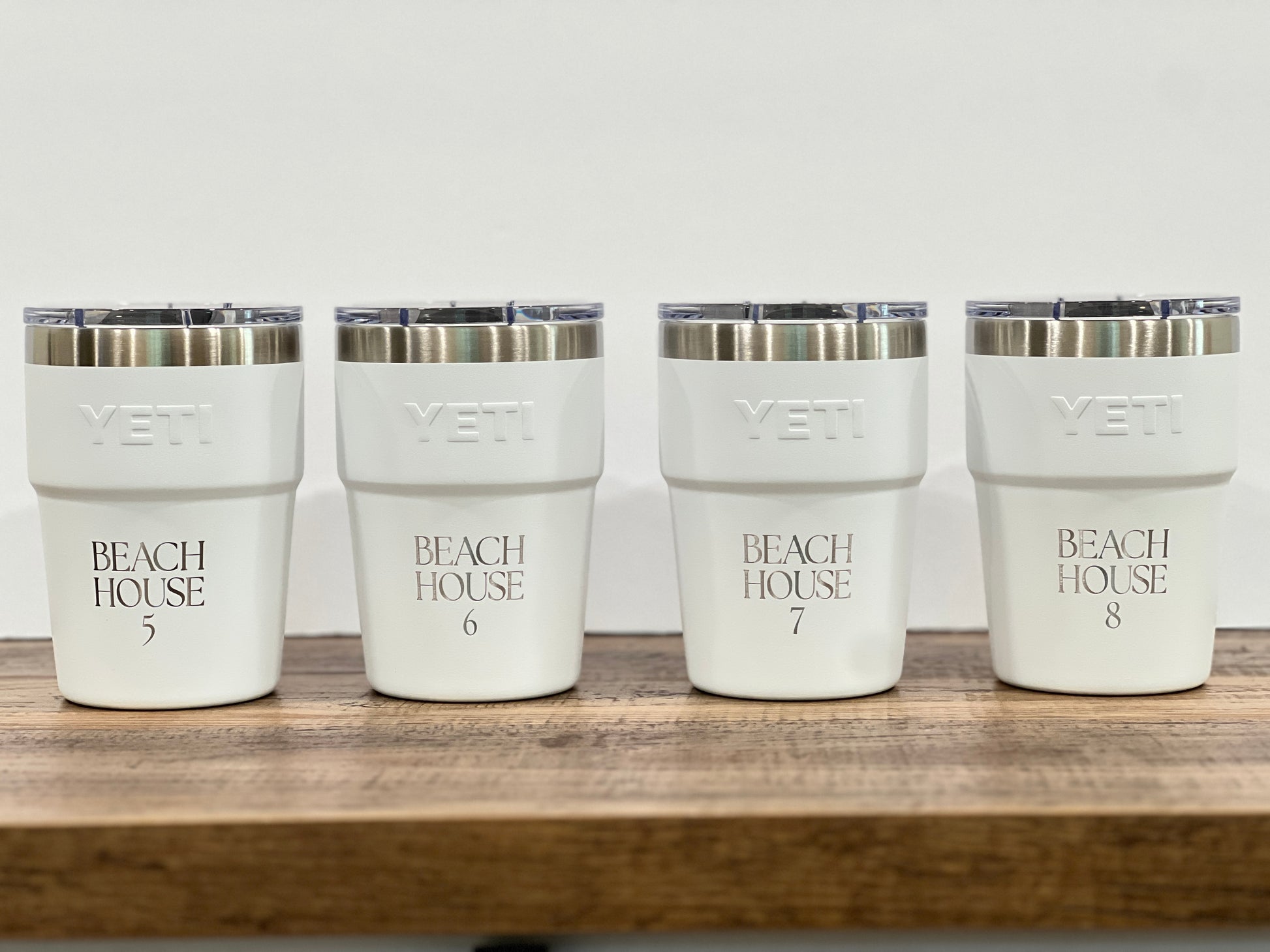 Four white YETI insulated tumblers with clear lids and silver rims, lined up on a wooden surface against a white background. Each cup is printed with 'BEACH HOUSE' and a number from 5 to 8.