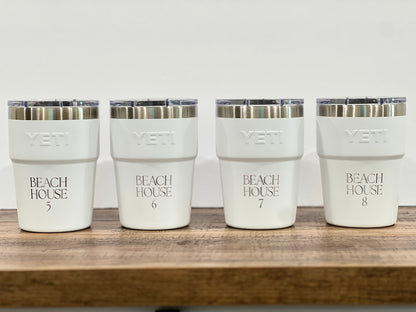 Four white YETI insulated tumblers with clear lids and silver rims, lined up on a wooden surface against a white background. Each cup is printed with 'BEACH HOUSE' and a number from 5 to 8.