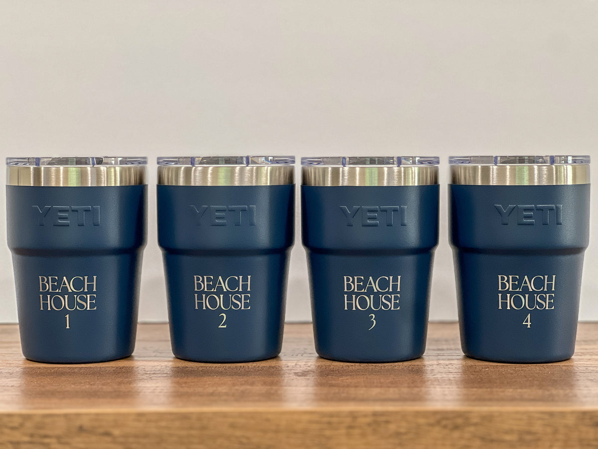 Four navy blue YETI insulated tumblers with clear lids and silver rims, arranged in a row on a wooden surface against a light beige background. The tumblers are personalized with 'BEACH HOUSE' and numbers 1 through 4.