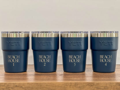 Four navy blue YETI insulated tumblers with clear lids and silver rims, arranged in a row on a wooden surface against a light beige background. The tumblers are personalized with 'BEACH HOUSE' and numbers 1 through 4.