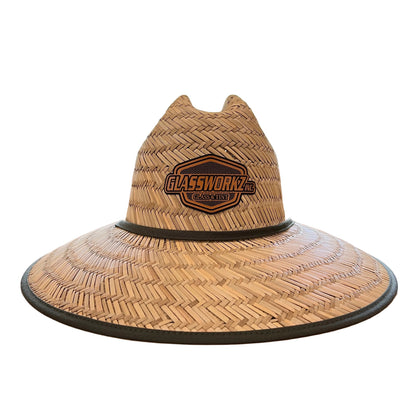 A woven straw cowboy hat with a black brim and a Harley-Davidson logo patch on the crown, displayed on a wooden table next to a window with blinds.