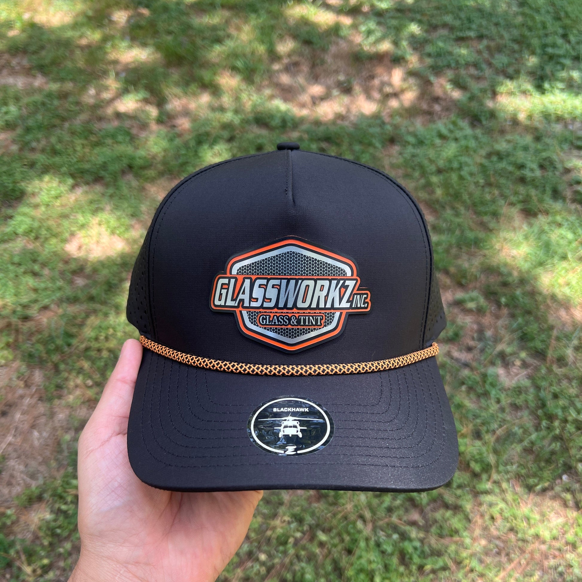 A black baseball cap held in a hand outdoors, featuring a large front patch that reads 'Glassworks' with a blue, orange, and white design, and a small circular logo on the side. The background is a blurred grassy outdoor area.
