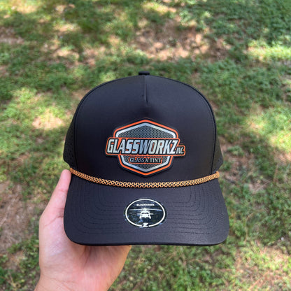 A black baseball cap held in a hand outdoors, featuring a large front patch that reads 'Glassworks' with a blue, orange, and white design, and a small circular logo on the side. The background is a blurred grassy outdoor area.
