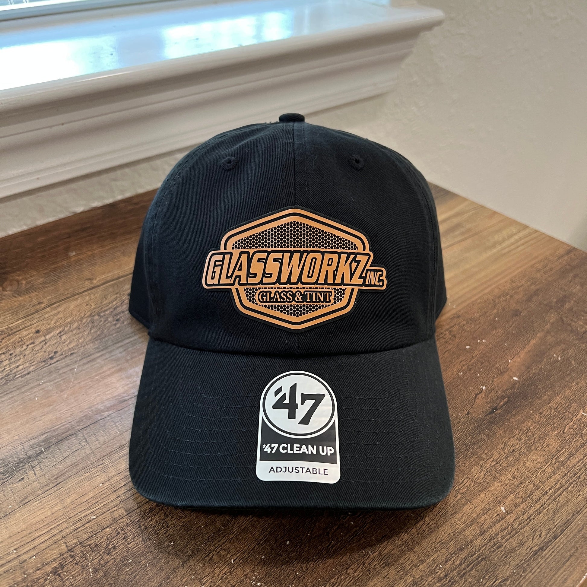 A black baseball cap resting on a wooden table, featuring a gold and black patch on the front and a white '47 My Clean Up Adjustable' sticker on the side.