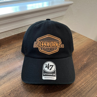 A black baseball cap resting on a wooden table, featuring a gold and black patch on the front and a white '47 My Clean Up Adjustable' sticker on the side.