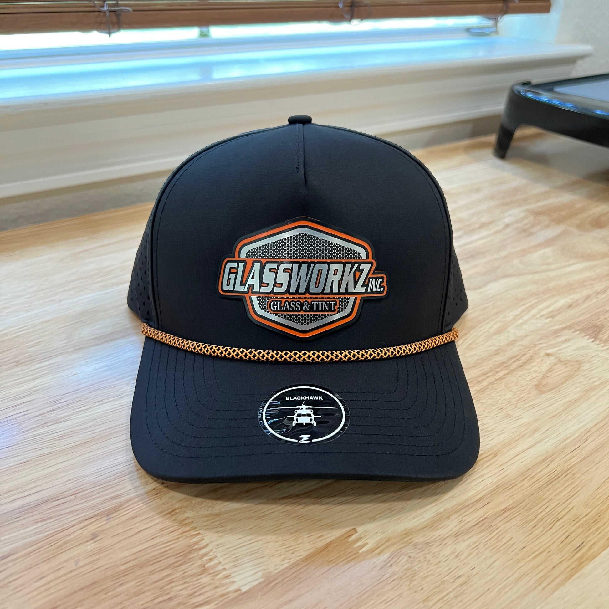 A black trucker-style cap with a gold chain detail and embroidered front patches, photographed on a light wood floor.