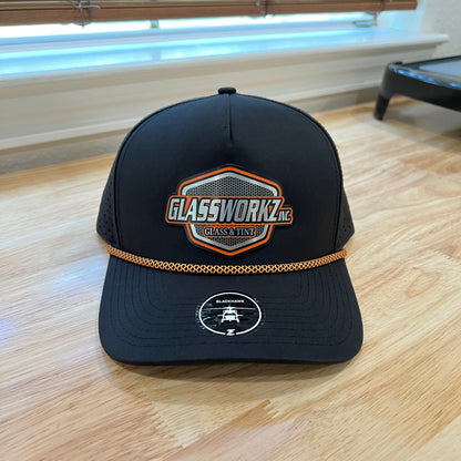 A black trucker-style cap with a gold chain detail and embroidered front patches, photographed on a light wood floor.