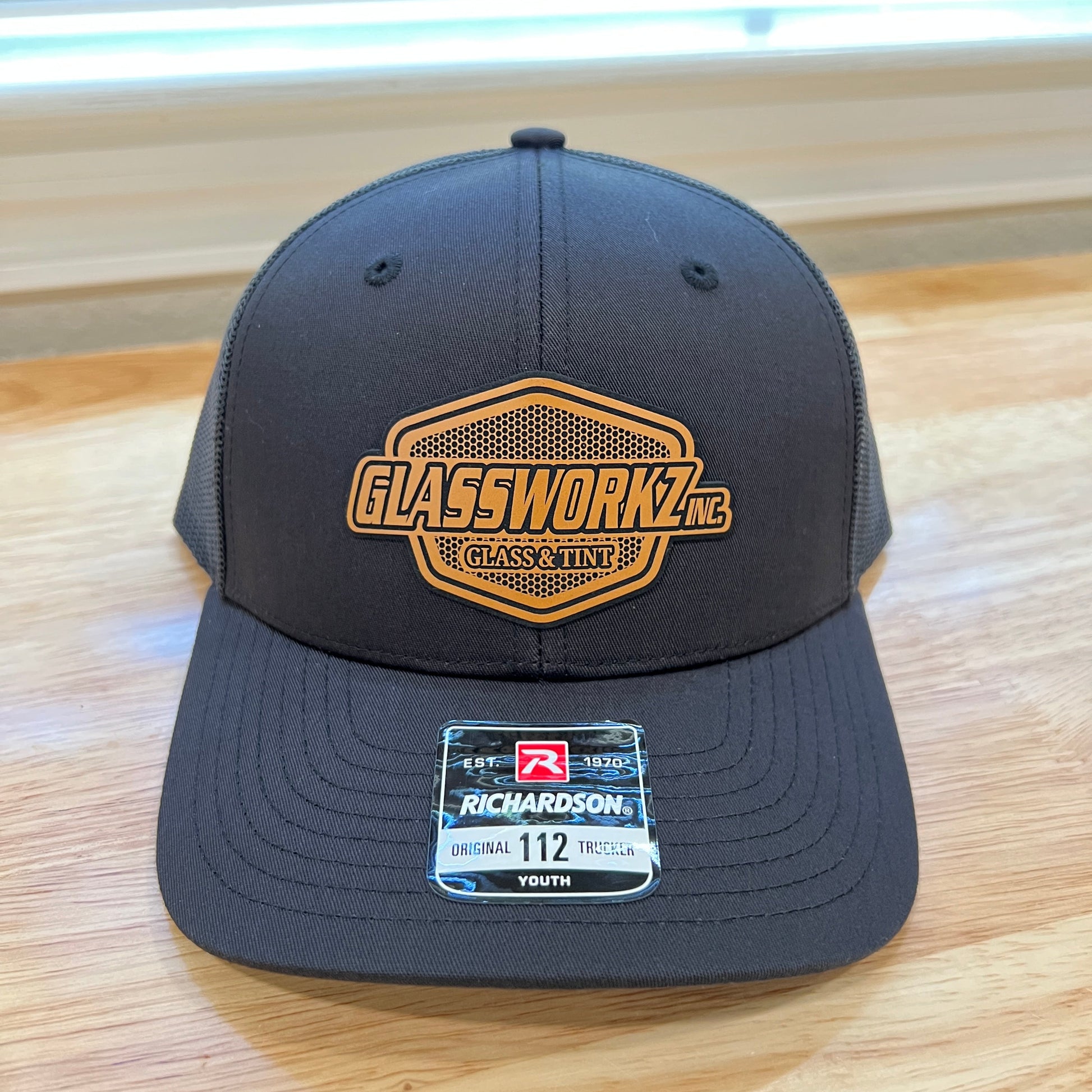 A dark gray trucker-style cap with a gold embroidered 'Glassworkz' logo on the front and a size 112 brand tag on the side, resting on a light wood floor.