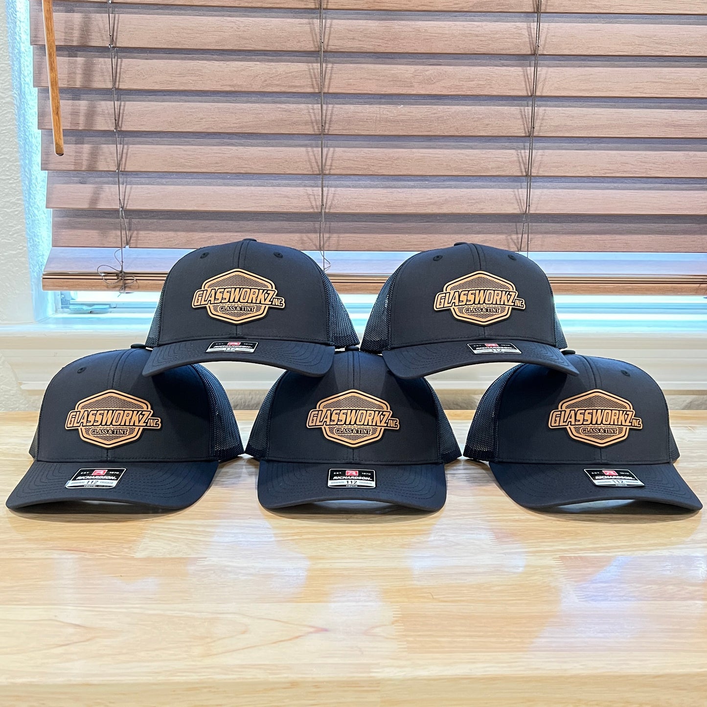 Six black baseball caps with a gold and white 'Allstar Works' patch on the front, arranged in two rows on a light wood table in front of window blinds.