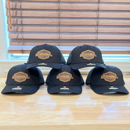 Six black baseball caps with a gold and white 'Allstar Works' patch on the front, arranged in two rows on a light wood table in front of window blinds.