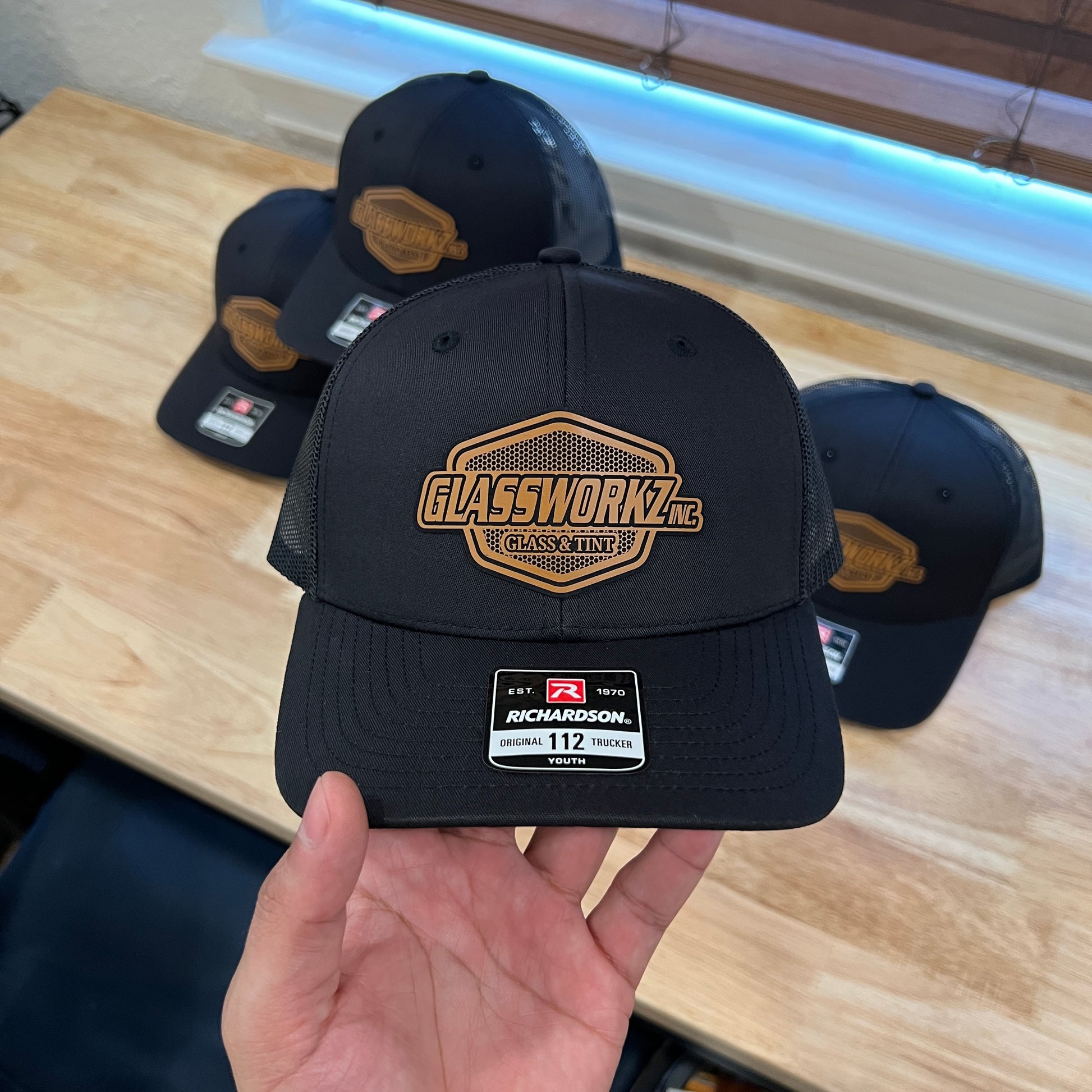 A hand holds a black Richardson 112 trucker hat with a gold and black 'GLASSWORKZ' patch on the front, with three more matching hats in the background on a wooden table.