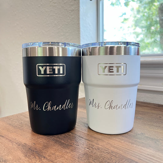 Two YETI tumblers, one white and one black, on a wooden surface with a window in the background.