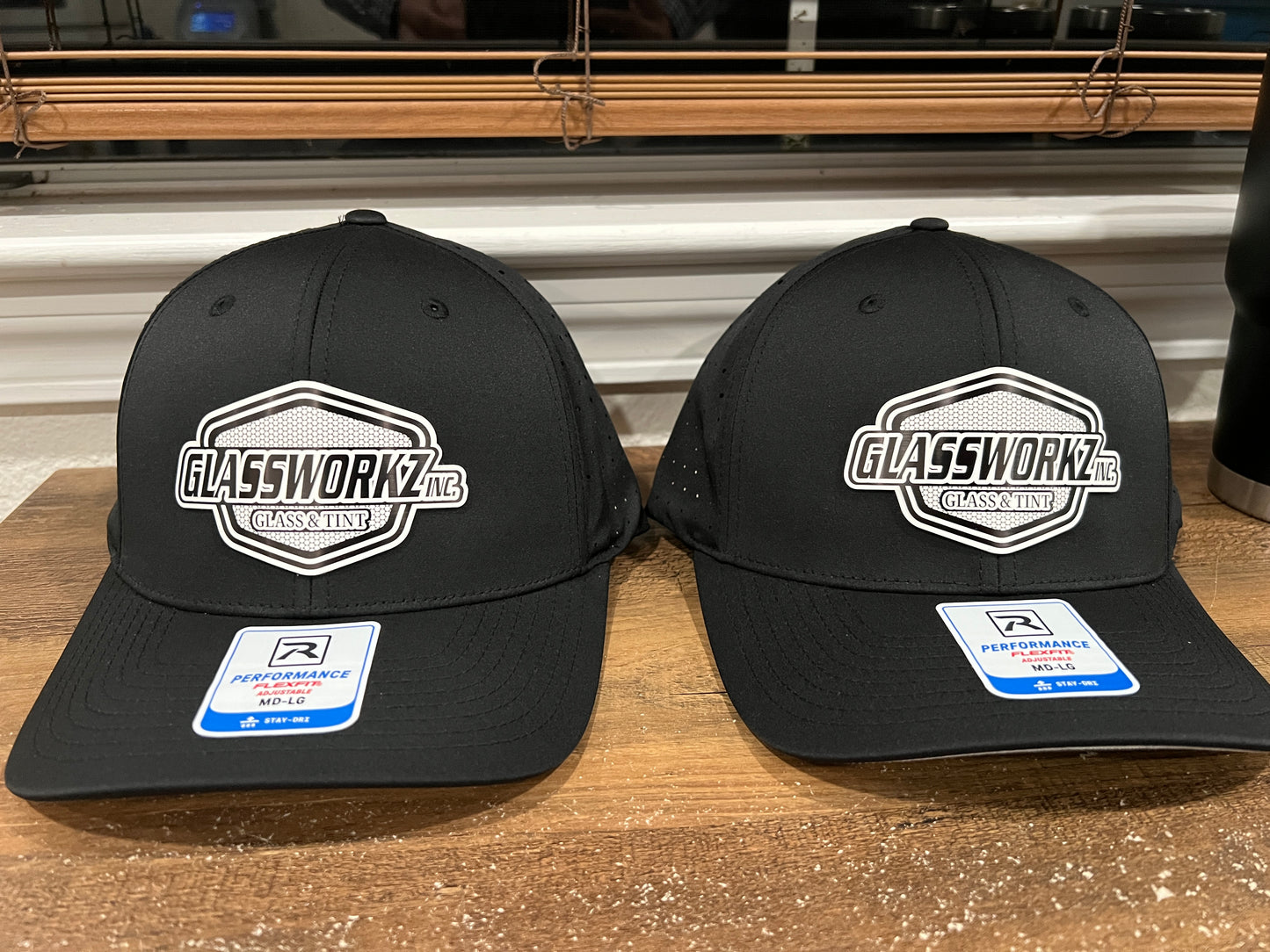 Two black structured baseball caps with white and gray 'GLASSWORKZ INC' logos on the front panels, displayed side-by-side on a wooden surface.