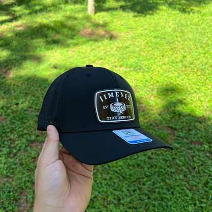 A black baseball cap with a white and black 'Jimenez' patch on the front, held by a hand against a green grass background.