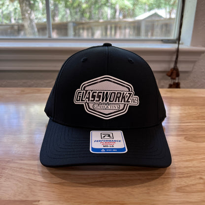 A black baseball cap with a white and gray 'GLASSWORKZ' logo on the front, resting on a light wood table in front of a window.