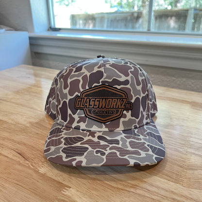 A camouflage baseball cap with a brown and gold 'GLASSWORKZ, INC.' patch on the front, resting on a light wood table in front of a window.