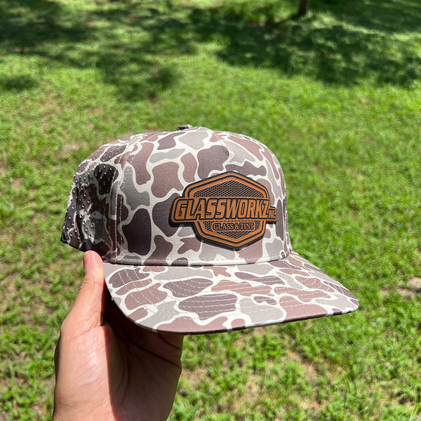 A hand holds a camouflage baseball cap outdoors in a grassy wooded area. The cap has a brown and gray camo pattern with a prominent brown embroidered patch on the front that reads 'GLASSWORKS CANVAS'.