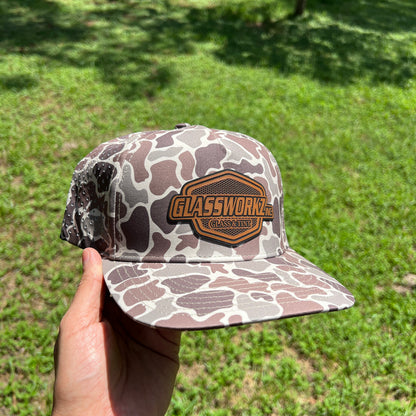 A hand holds a camouflage baseball cap outdoors in a grassy wooded area. The cap has a brown and gray camo pattern with a prominent brown embroidered patch on the front that reads 'GLASSWORKS CANVAS'.