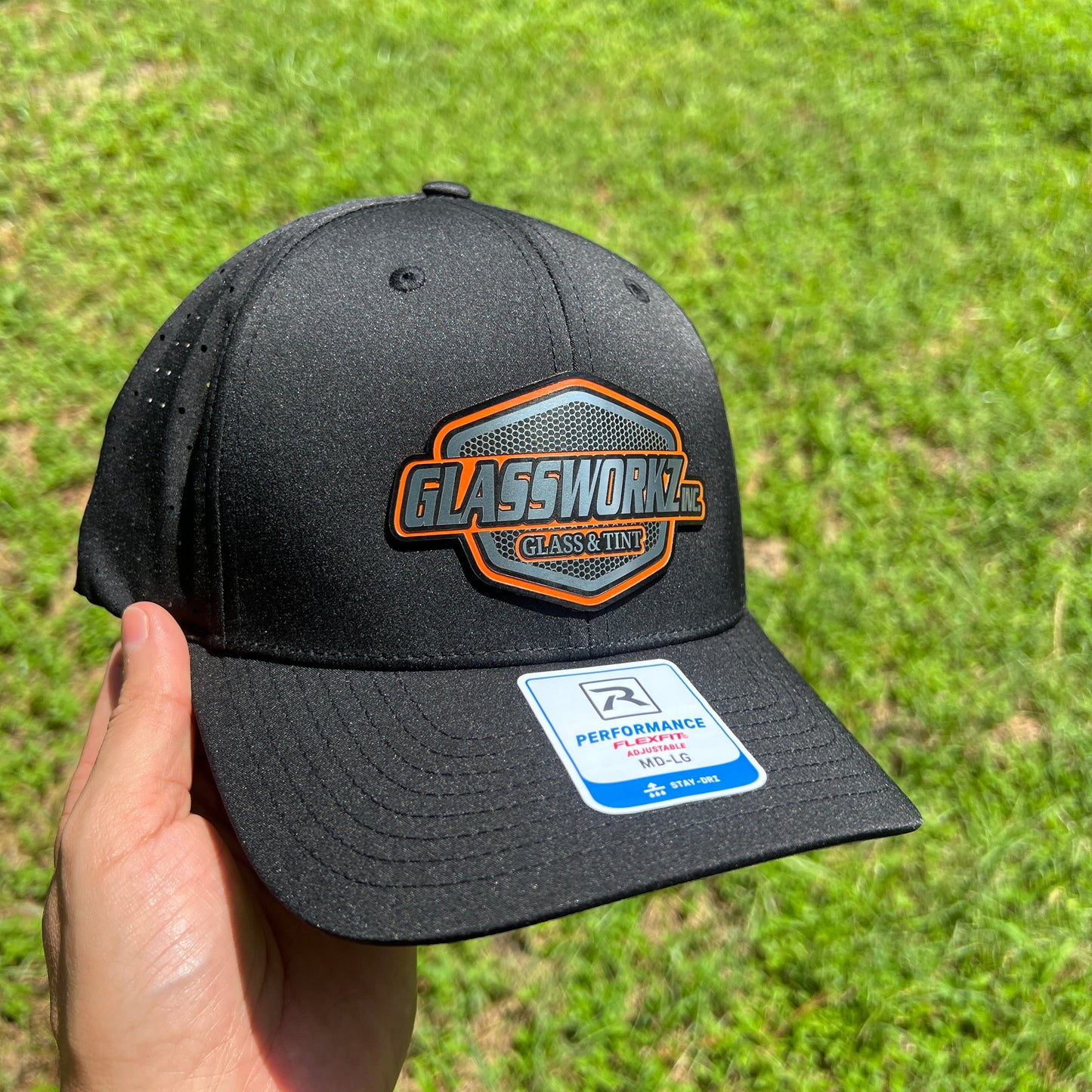 A black baseball cap with a orange and black patch on the front and a white sticker on the brim, held by a hand against a green grass background.