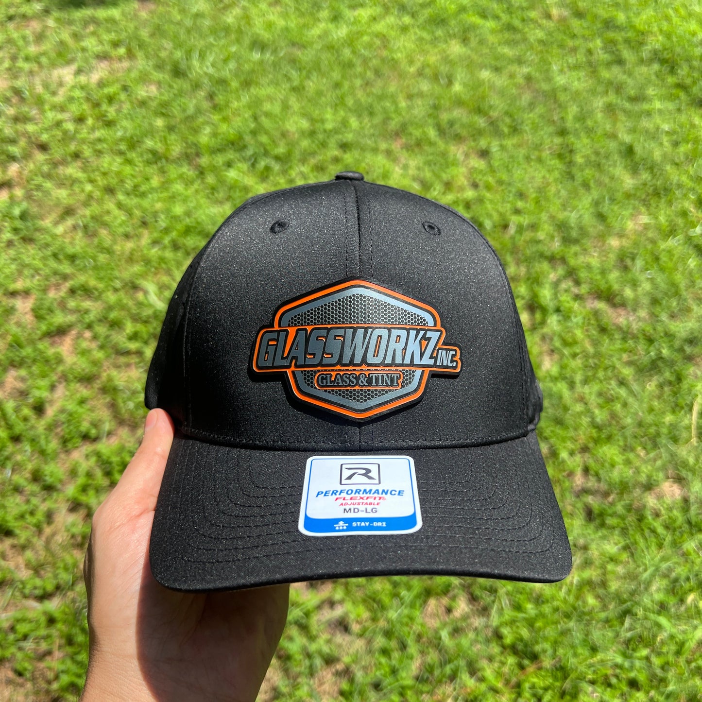 A black baseball cap held by a hand against a green grass background. The cap features a prominent hexagonal patch on the front with the text 'GLASSWORKZ' and 'CLASSIC CAP' in orange and white, along with a blue and white 'Performance Fit' sticker on the brim.